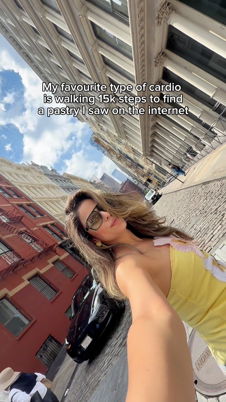 Roopsi Bansal taking a selfie on a downtown NYC street lined with a red brick building and fire escapes, wearing a yellow floral top