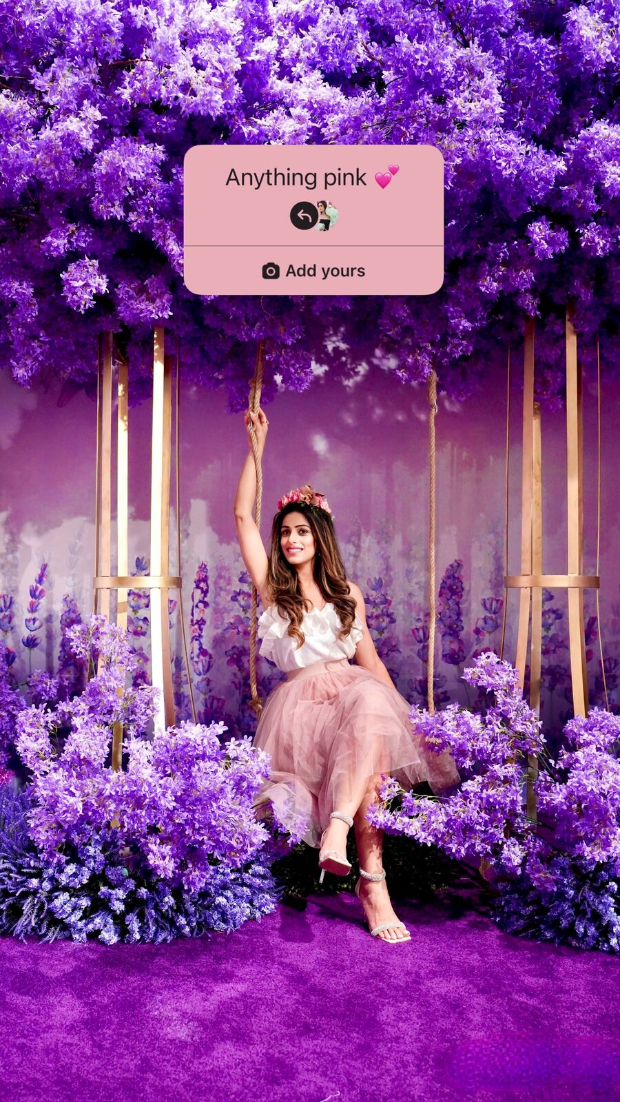 Roopsi Bansal in a pink tulle skirt and floral crown on a swing surrounded by purple flowers, with an 'Anything pink' Add Yours sticker overlay