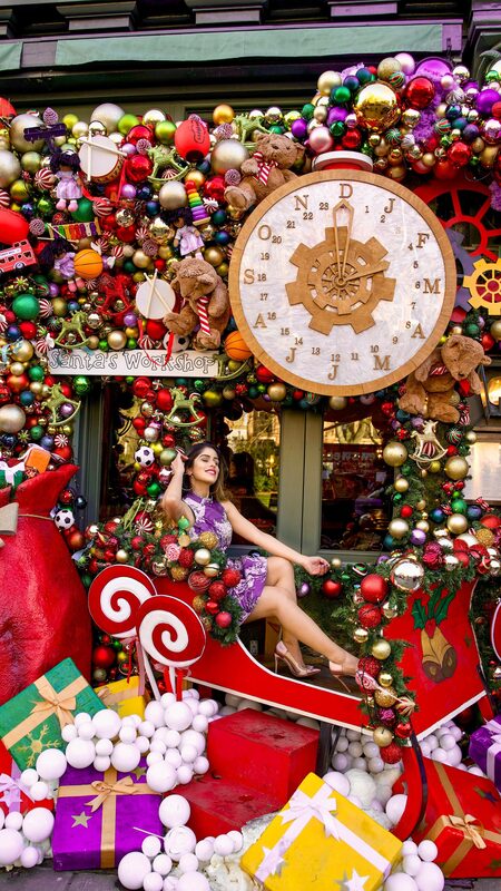 Roopsi Bansal in a white embroidered midi dress, holding a neon green handbag, standing inside a giant walk-through Christmas ornament sculpture with palm trees behind her