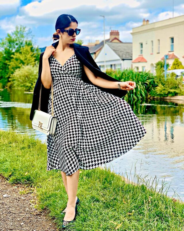 Standing by a pond with a black blazer draped over the shoulders of a black and white houndstooth midi dress, sunglasses, and a white Ralph Lauren bag