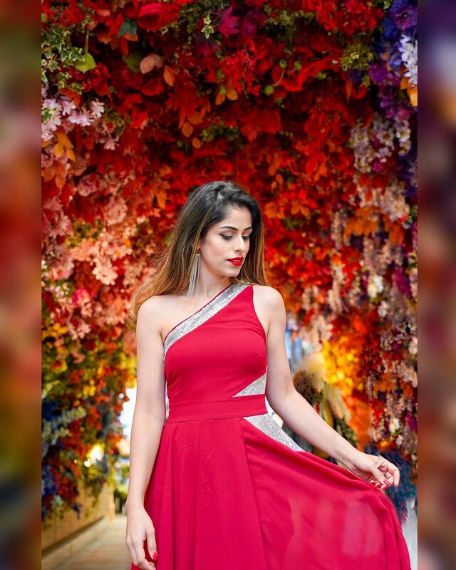 Roopsi Bansal in a red one-shoulder dress with a silver diagonal trim, standing in a tunnel of vivid red and purple flowers