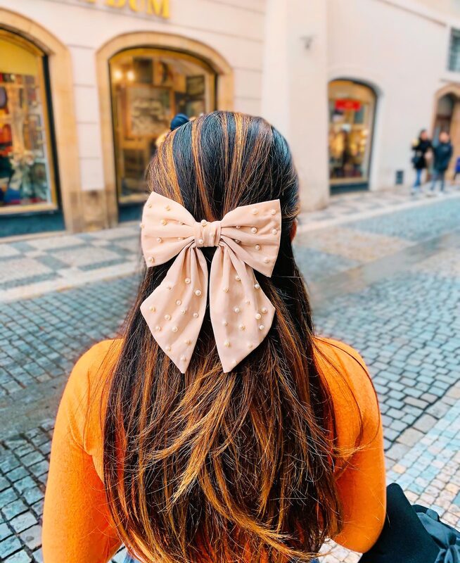 Back-of-head view of a large pearl-studded bow hair clip in half-up hair, wearing an orange sweater, on a Prague cobblestone street lined with shop windows