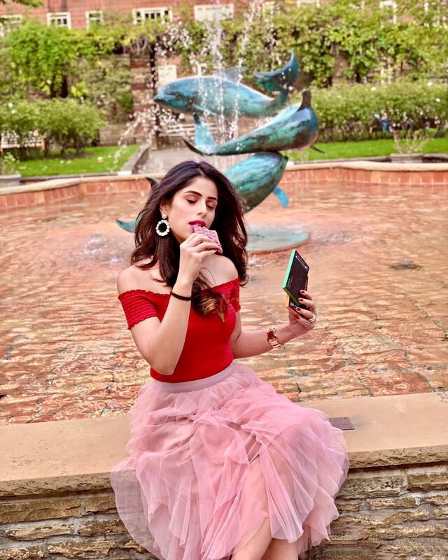 Roopsi Bansal in a red top and pink tulle skirt, tasting a diamond-shaped chocolate bar in front of a dolphin fountain
