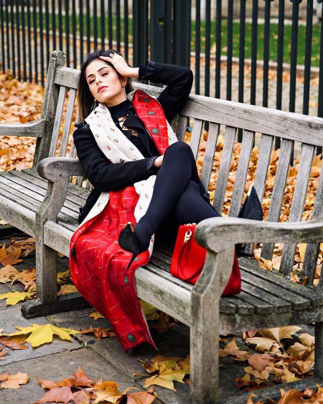 Roopsi Bansal in a black coat with a long red plaid scarf and cream floral scarf, reclining on a park bench surrounded by fallen autumn leaves