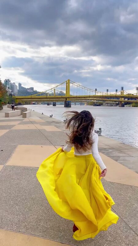 Roopsi Bansal from behind in a bright yellow maxi skirt and white knit top, hair blowing in the wind, walking along the riverfront near the yellow Roberto Clemente Bridge in Pittsburgh