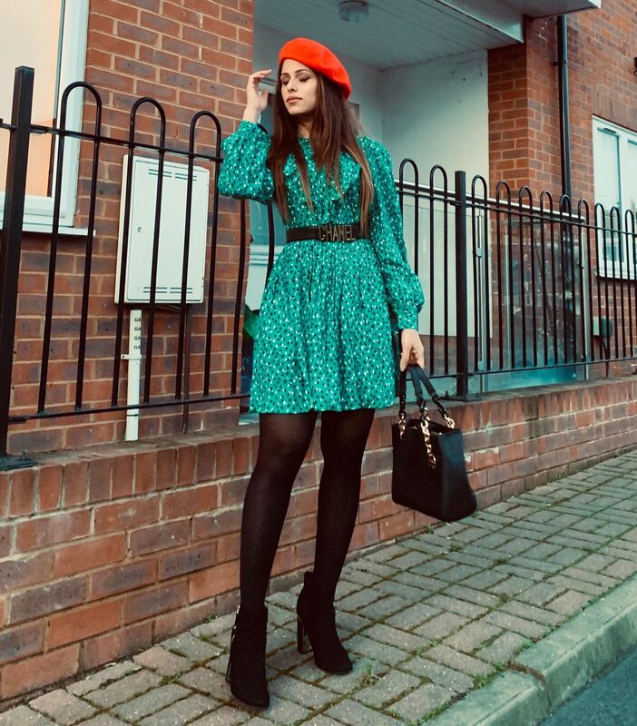Standing against a brick building with black railings in Lyon, France, wearing a red beret, a green polka-dot smock dress with a Chanel logo belt, black tights, and ankle boots