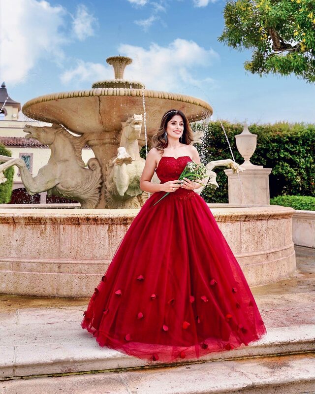 Roopsi Bansal in a strapless red ball gown scattered with fabric rose petals, holding a small bouquet of white flowers in front of a stone fountain with sculpted seahorses