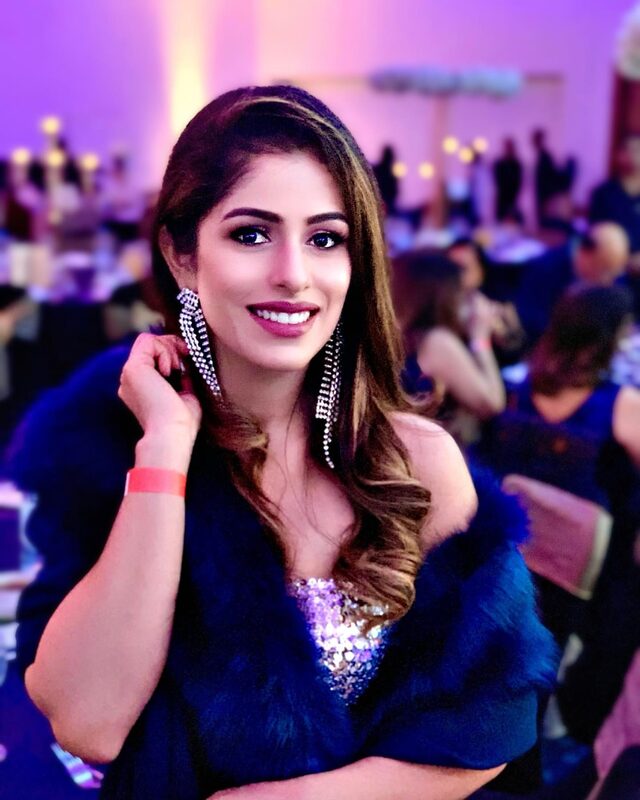 Smiling portrait at an evening event, wearing a navy sequined top and blue faux-fur wrap, sparkly fringe drop earrings, under colorful purple and gold event lighting