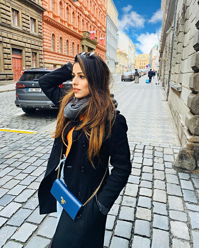 Standing on a Prague street looking off to the side, wearing a black coat over an orange top, grey scarf, sunglasses pushed up, and a blue Prada crossbody bag