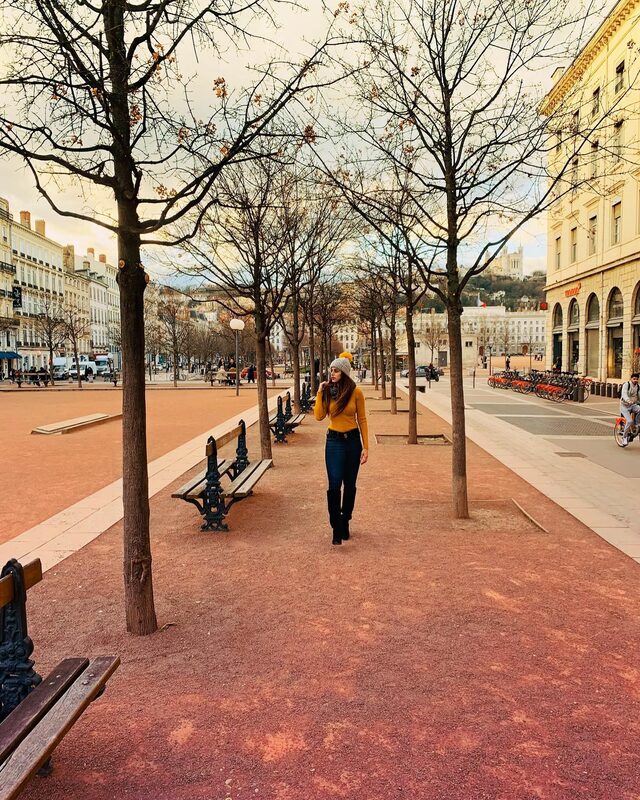 Walking through a wide, tree-lined square past park benches, wearing a mustard sweater, grey pom-pom beanie, and grey scarf, jeans, and black boots