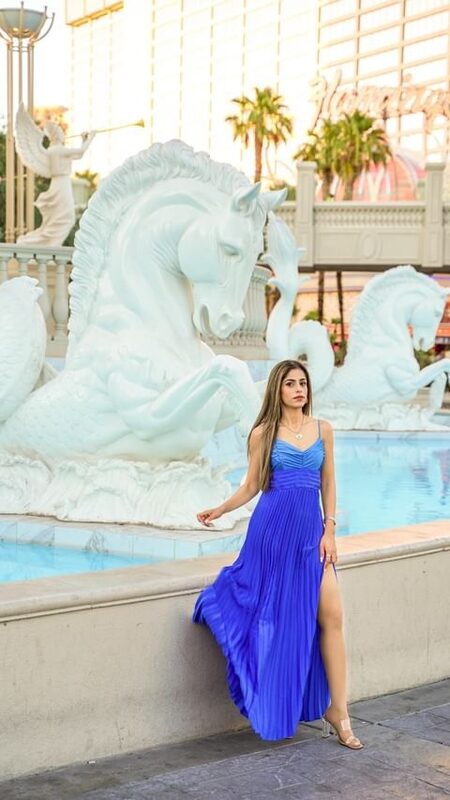 Roopsi Bansal in a cobalt blue pleated gown leaning against the ledge at the Bellagio fountains in Las Vegas, with marble seahorse sculptures and the Strip behind her