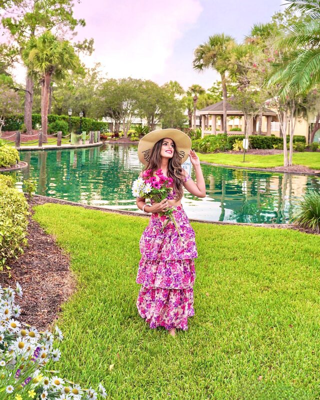 Roopsi Bansal in a pink and purple floral tiered dress and a wide straw hat, holding a bouquet of daisies beside a lagoon at Wyndham Orlando Resort