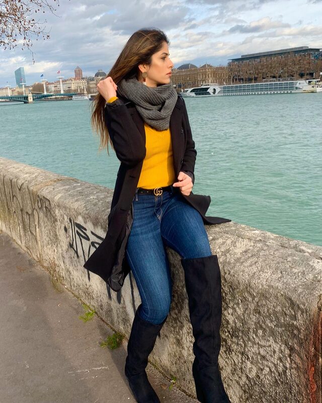 Sitting on a stone riverside wall in Lyon, France, wearing a mustard sweater, black coat, grey scarf, jeans with a Gucci belt, and over-the-knee black suede boots, with the river and city skyline behind