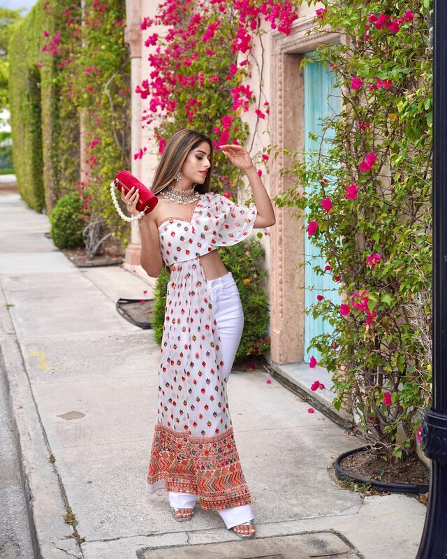 Roopsi Bansal in a white one-shoulder embroidered fusion top and white pants, holding a red clutch, beside a bougainvillea-covered wall and a turquoise door