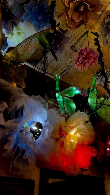 A ceiling covered in giant flowers, glowing masks, and a green parrot ornament at an Alice in Wonderland themed restaurant
