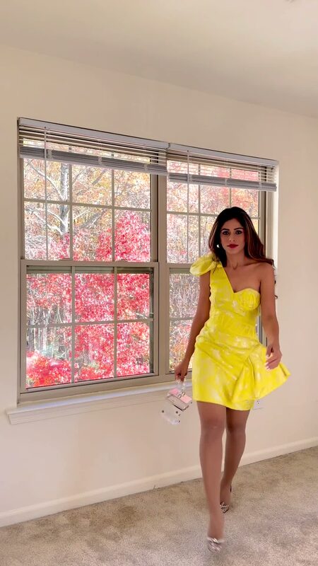 Roopsi Bansal in a neon yellow one-shoulder mini dress indoors, red fall foliage visible through the window behind her