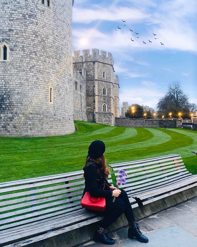 Sitting on a park bench facing away from the camera, wearing a black beret and all black outfit, holding purple flowers and a red bag, looking toward Windsor Castle's stone towers and manicured lawns