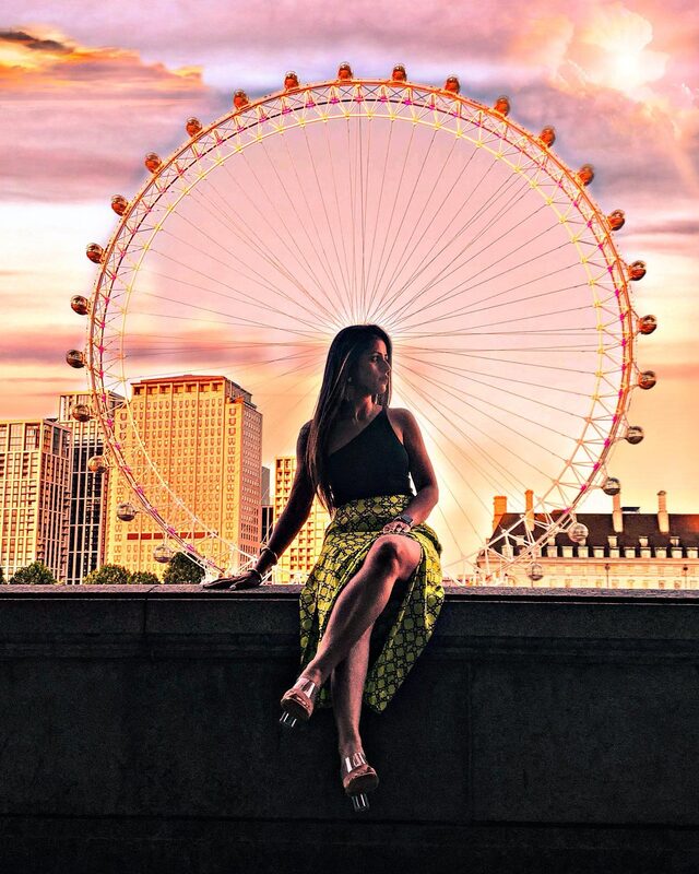 Silhouetted against a golden-pink sunset with the London Eye lit up behind her, wearing a black one-shoulder top and a yellow patterned wrap skirt