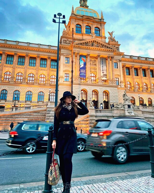 Standing across a busy street from Prague's National Museum at dusk, wearing a black coat with a Chanel logo belt, leopard-print scarf, black hat, and a Burberry check bag, with traffic passing in the foreground
