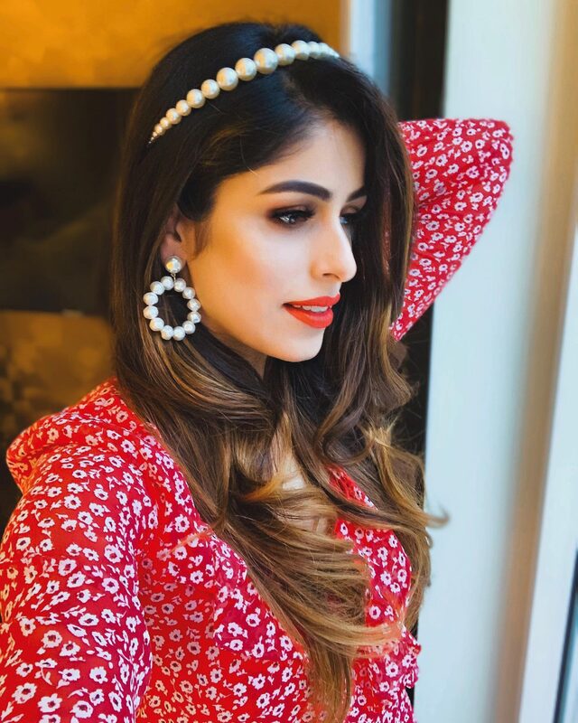 A close-up portrait in a red floral wrap dress and pearl headband, with warm makeup and a bold red lip, sitting on a bed in a dimly lit hotel room
