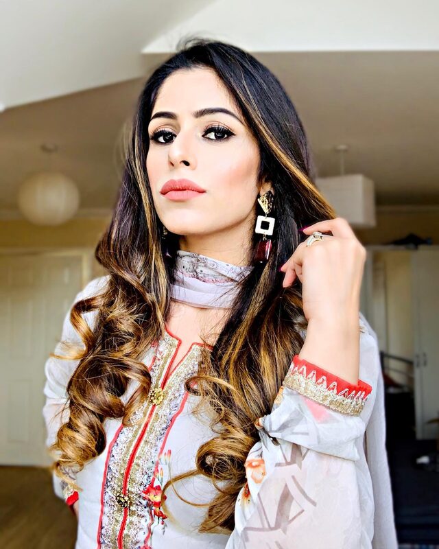 A close-up portrait wearing a white and gold embroidered Indian kurta with red trim, statement drop earrings, and curled hair
