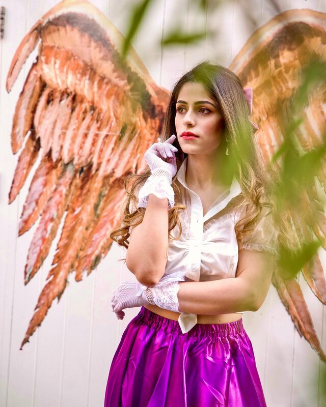 Roopsi Bansal in a white satin blouse, magenta skirt, and white lace gloves, standing in front of a mural of painted angel wings at Neal's Yard, London