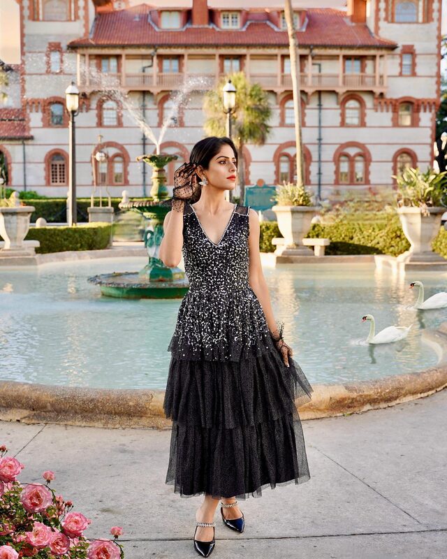 Roopsi Bansal in a black sequined tiered dress and sheer polka-dot gloves, standing beside a fountain with swans in front of the ornate Casa Monica hotel building