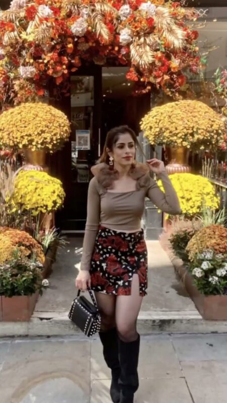 Roopsi Bansal in a brown ruffle-sleeve top and floral mini skirt, posing in front of a floral entrance archway with chrysanthemum planters