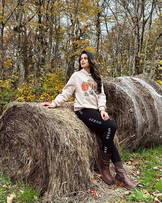 Roopsi Bansal in a cream pumpkin-print hoodie sitting on hay bales in an autumn forest