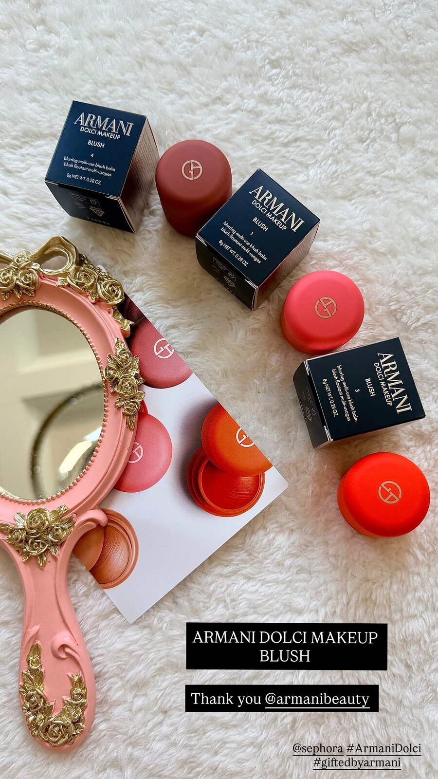 Four Armani Beauty Dolci Makeup blush balms with their boxes and a rose-gold hand mirror on a white shag rug
