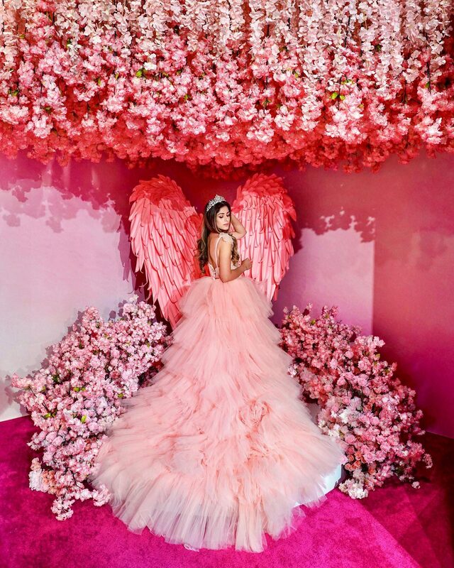 Roopsi Bansal in a pink tulle ballgown with feathered angel wings and a tiara, under a canopy of pink flowers at a pink-themed installation