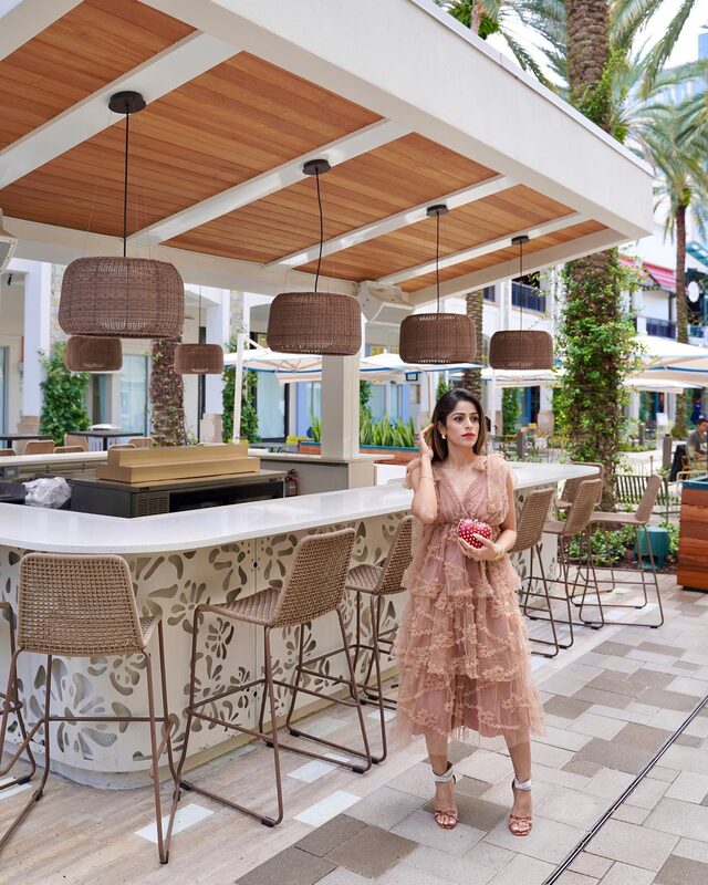 Roopsi Bansal in a blush pink tiered ruffle dress, holding a red heart-shaped clutch, standing at an outdoor poolside bar under woven pendant lights