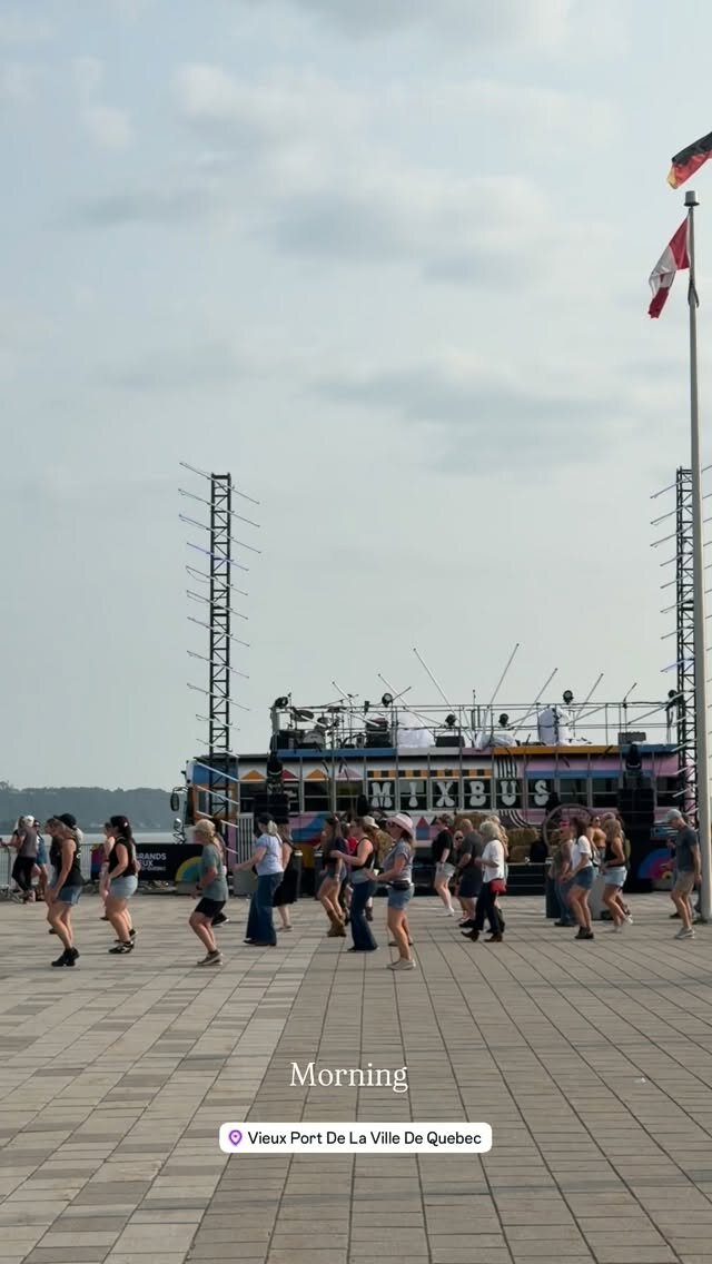 A crowd of people line dancing on the waterfront plaza at Vieux Port de Quebec in front of a colorful mobile stage, with Canadian and German flags flying nearby