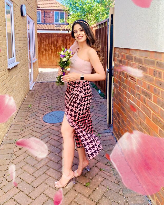 Roopsi Bansal in a blush one-shoulder top and a pink-and-black houndstooth wrap skirt with a thigh slit, holding a bouquet of flowers
