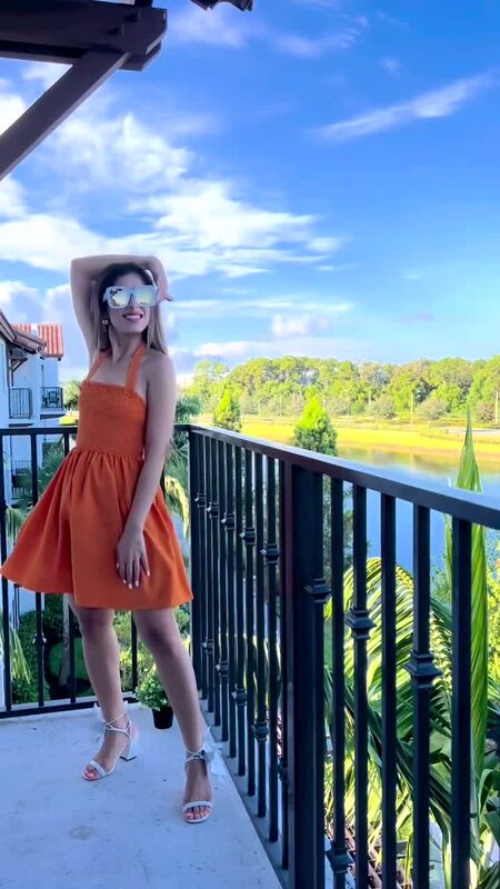 Roopsi Bansal in a bright orange halter dress on a balcony overlooking a lake