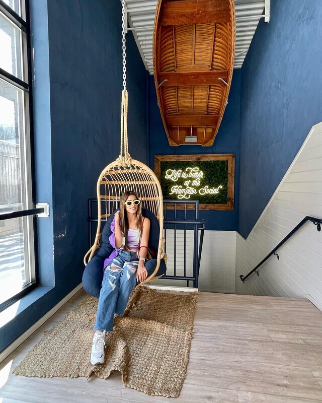 Roopsi Bansal sitting in a hanging rattan swing chair at The Hampton Social, wearing a purple tie-dye crop top and ripped jeans, with a neon sign reading 'Life is better at the Hampton Social' behind her