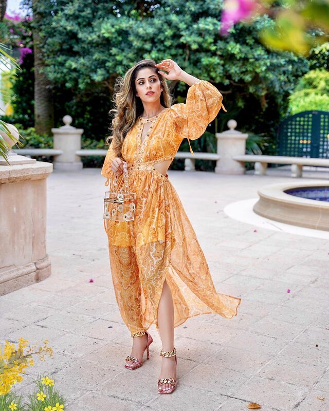 Roopsi Bansal in a marigold paisley-print maxi dress with waist cutouts, holding a clear floral box clutch, walking past a garden fountain in West Palm Beach