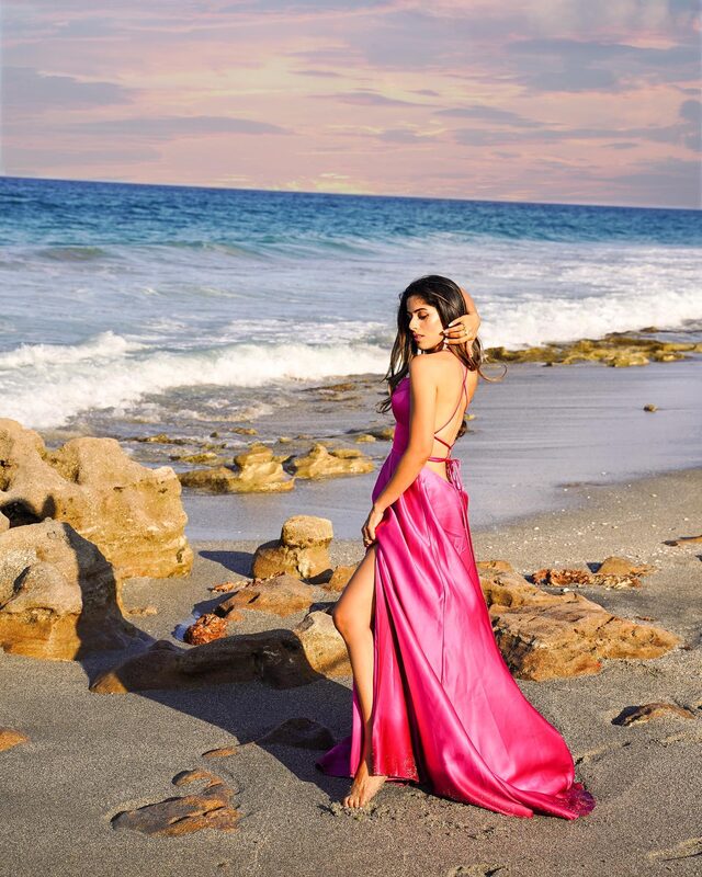 Roopsi Bansal in a fuchsia satin gown with a thigh slit, standing on the rocky shoreline of Coral Cove Park, Florida at golden hour