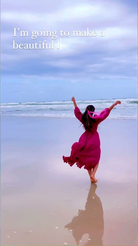 Roopsi Bansal in a flowing magenta dress from Chicwish, both arms raised, walking barefoot on the sand at the water's edge