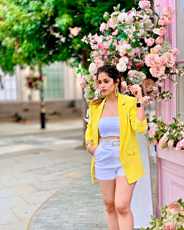 Roopsi Bansal in a yellow blazer and white co-ord, standing in front of the flower-covered facade of Peggy Porschen's cafe in London