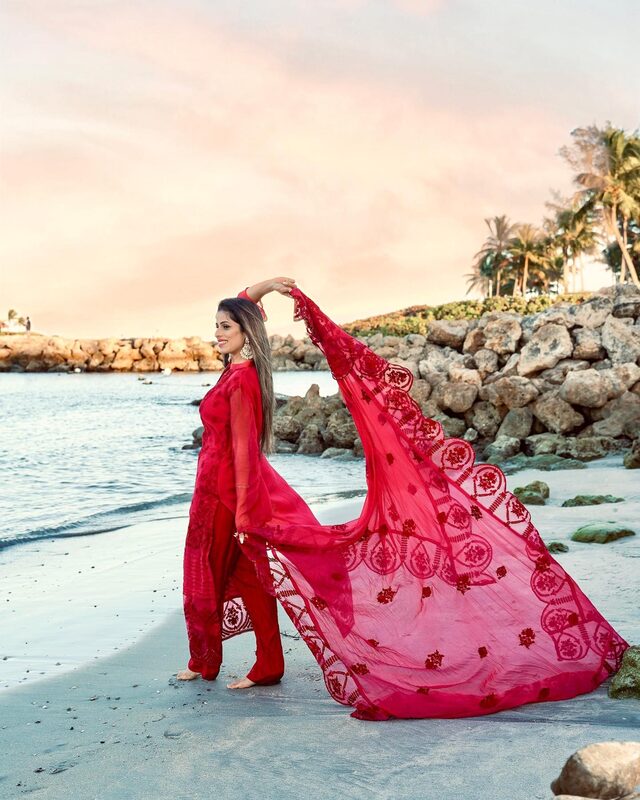 Roopsi Bansal in a red printed kurta suit, holding up a matching sheer red dupatta that billows in the wind, standing on a rocky shoreline at Dubois Park at sunset