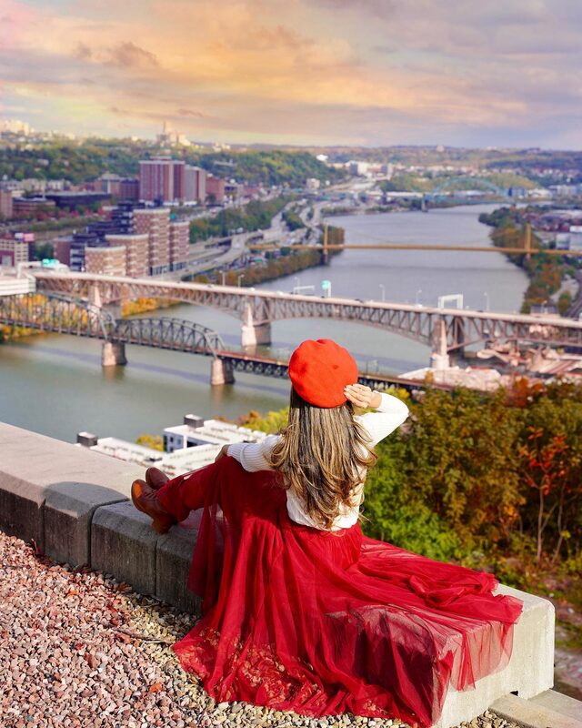 Roopsi Bansal from behind in a red tulle skirt and red beret, seated on a stone ledge overlooking downtown Pittsburgh and its bridges from a hillside lookout