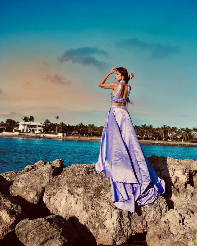 Roopsi Bansal in a beaded lilac open-back crop top and flowing lilac satin skirt, standing on coral rocks at Dubois Park with the ocean behind her