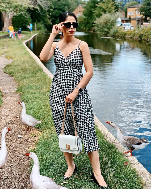 Standing on the grass beside a canal surrounded by geese, wearing a black and white houndstooth sundress, sunglasses, and holding a white River Island bag