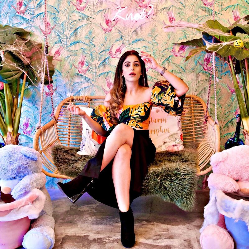 Roopsi Bansal in a tropical print off-shoulder top and black skirt, sitting on a rattan swing chair between two large teddy bears at a candy cafe
