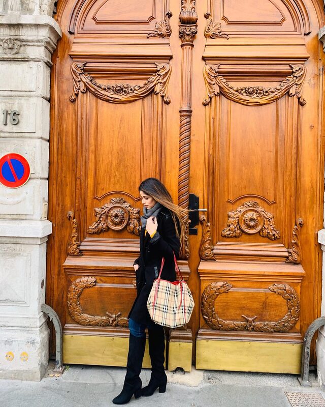 A side profile view standing before the same carved wooden doors in Lyon, scarf catching the wind
