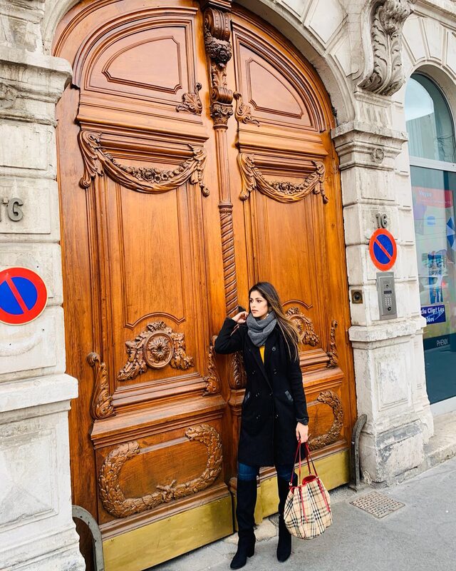 Standing before a massive ornately carved wooden double door in Lyon, France, wearing a black coat, grey scarf, jeans, over-the-knee boots, and carrying a Burberry check bucket bag
