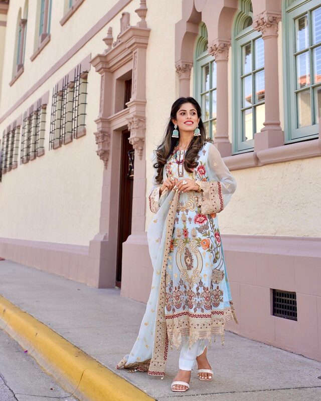 Roopsi Bansal in a pastel blue embroidered salwar suit with a floral-embroidered dupatta from Khas, standing in front of an ornate Mediterranean Revival building with green-shuttered windows