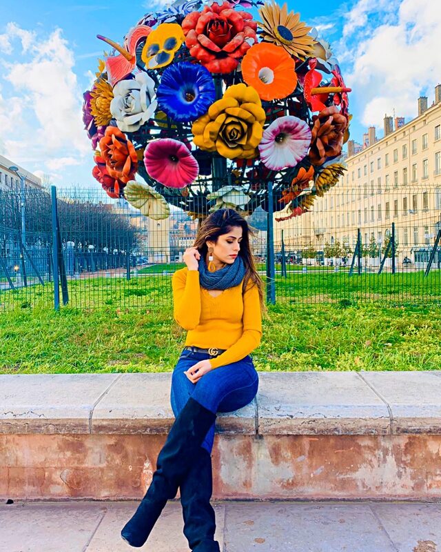 Sitting on a low wall beneath a giant sculptural bouquet made of painted metal flowers, wearing a mustard sweater, grey scarf, and jeans with a Gucci belt, green lawn and buildings behind