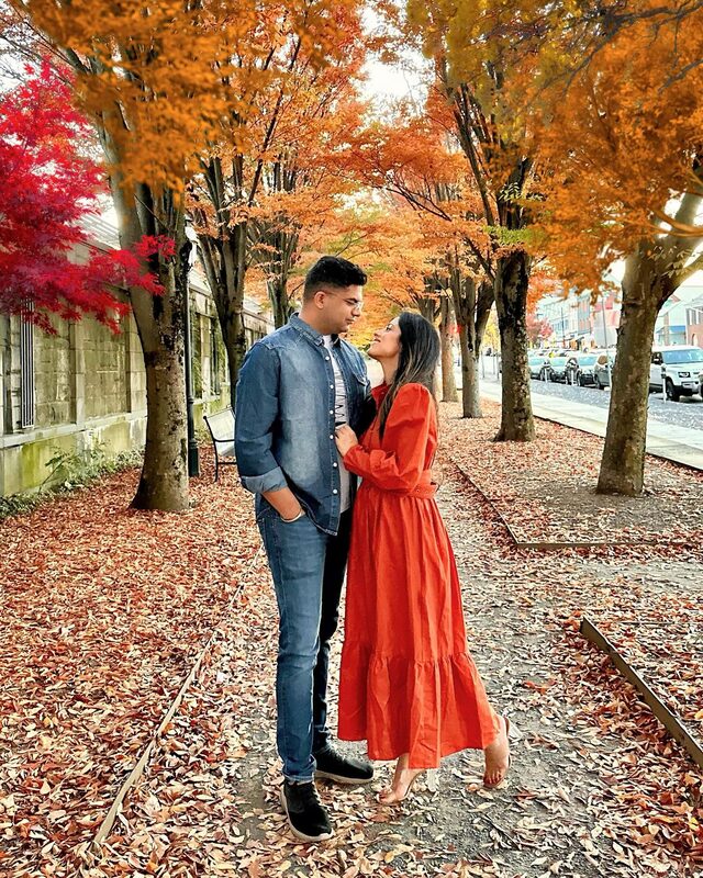 Roopsi Bansal in a rust-orange dress from Chicwish and her husband walking under a canopy of fall foliage